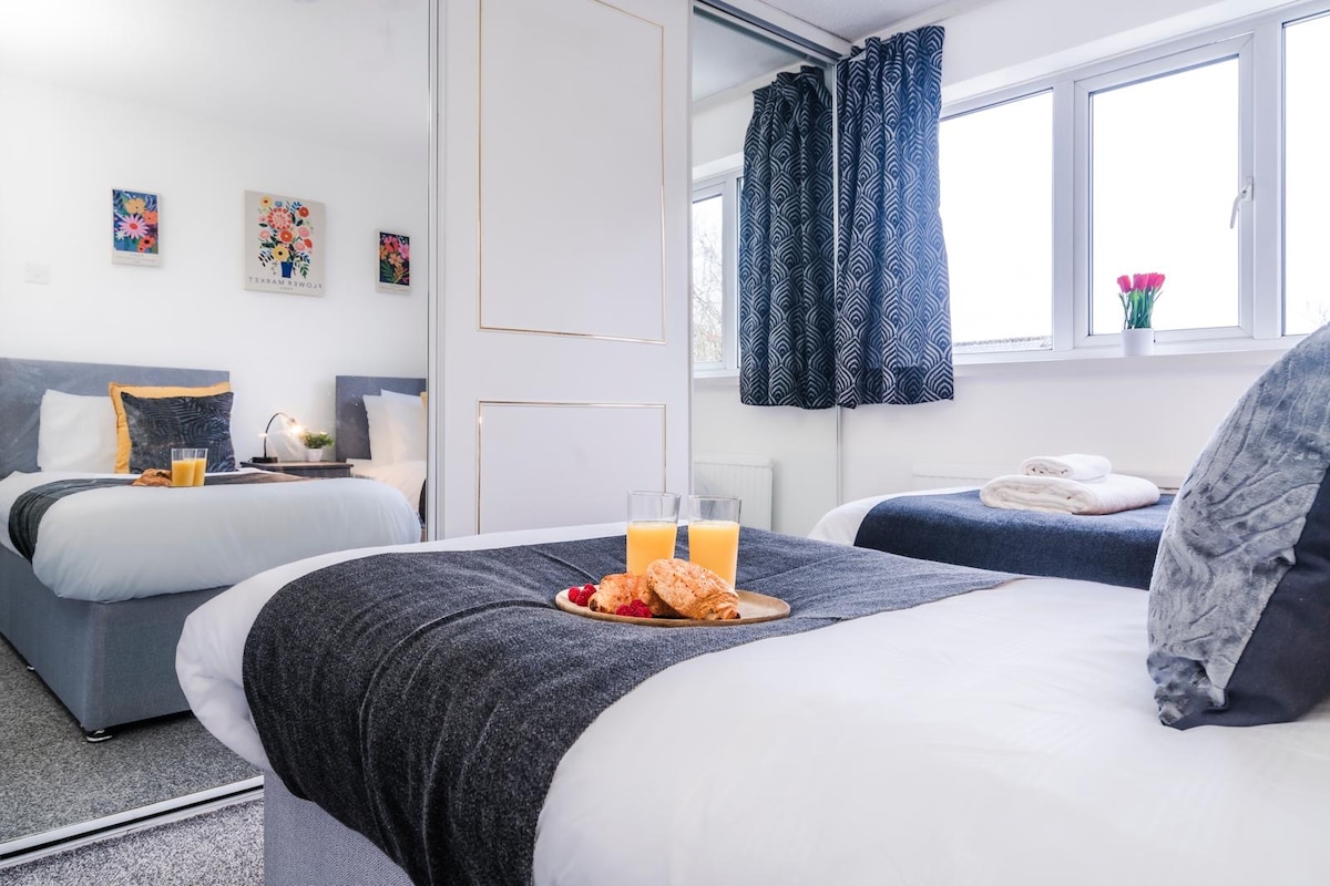 The bedroom showcases a spacious bed adorned with a dark coverlet, complemented by decorative pillows. A tray with orange juice and pastries is placed at the foot of the bed. Natural light filters through multiple windows, enhancing the bright and airy atmosphere.