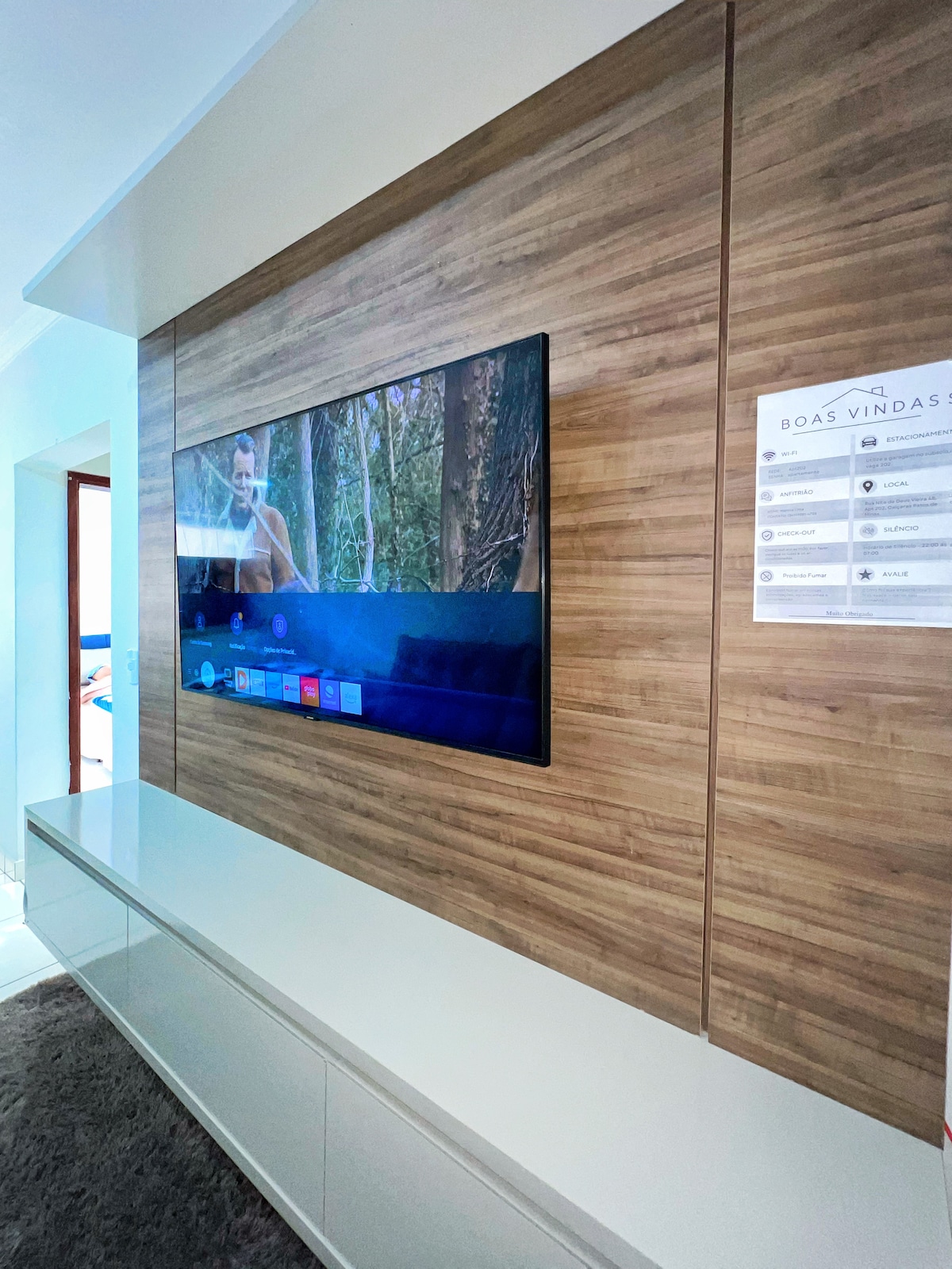 A sleek entertainment area features a large, flat-screen TV mounted on a wooden accent wall. Below, a modern white media console provides storage. Natural light is visible in the background, enhancing the space's inviting look.