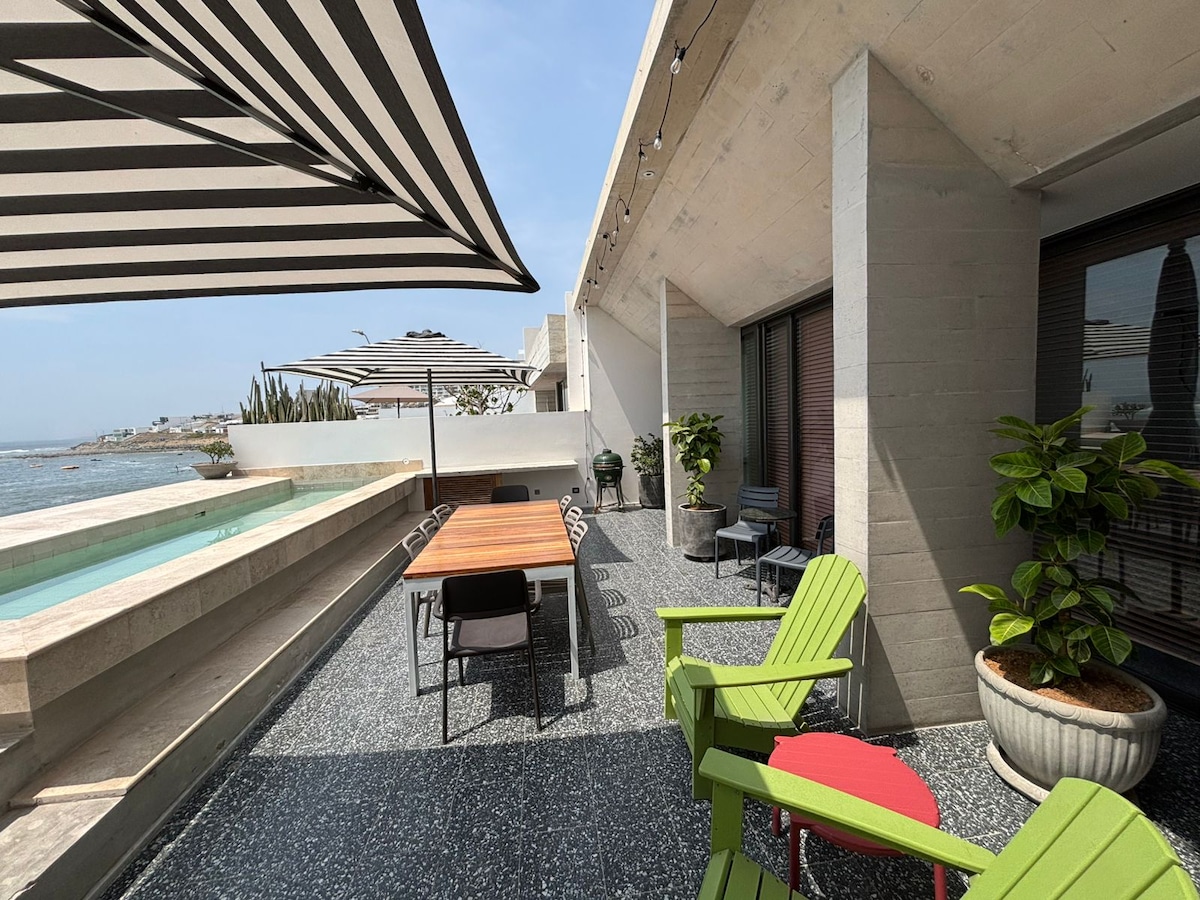 A spacious terrace overlooks the ocean, featuring a rectangular swimming pool and multiple outdoor seating areas. A wooden dining table is paired with contemporary chairs, while vibrant green lounge chairs and a red accent chair add a pop of color. Shade is provided by striped awnings.