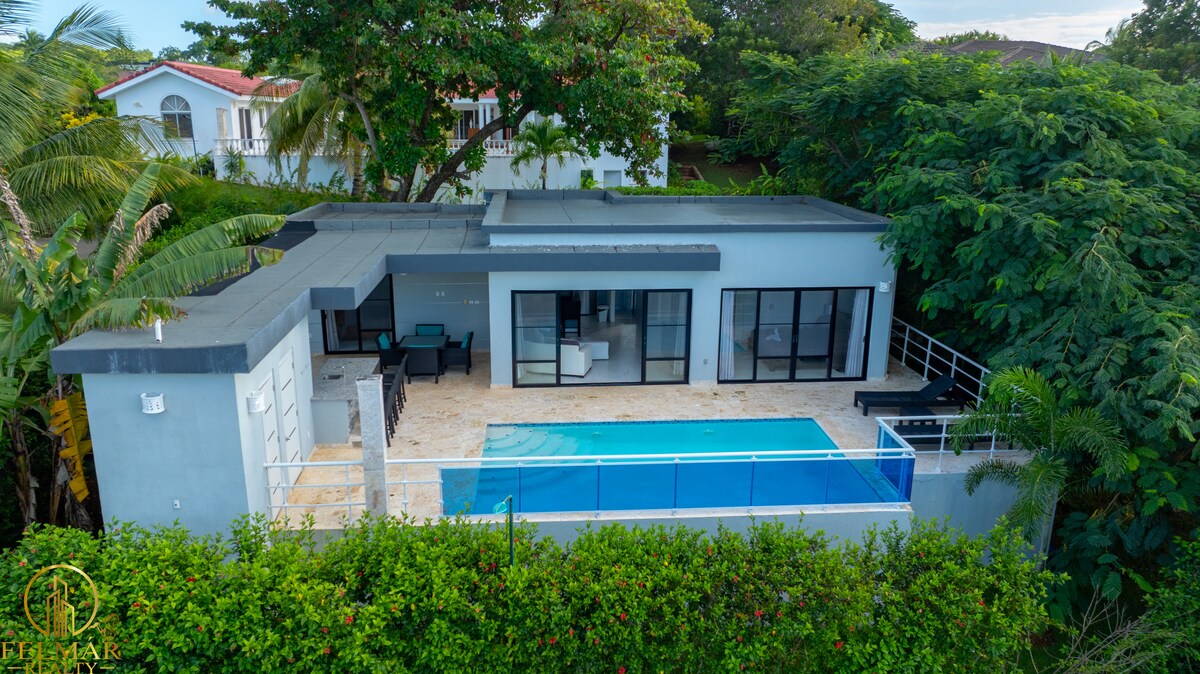 A modern house is set amid lush greenery, featuring a spacious terrace with a glistening pool. Floor-to-ceiling windows enhance the connection to the outdoors, while a minimalist seating area is visible on the terrace, blending seamlessly with the overall design.