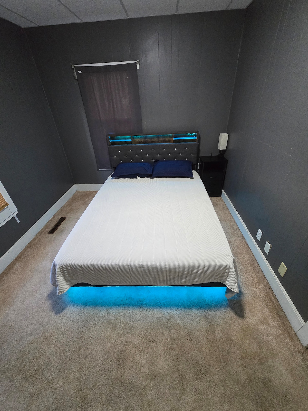 A neatly arranged sleeping area features a spacious bed with a light-colored coverlet and blue accent pillows. Soft carpet lines the floor, and a nightstand is positioned beside the bed. A subtle blue light shines beneath the bed, enhancing the soothing ambiance.