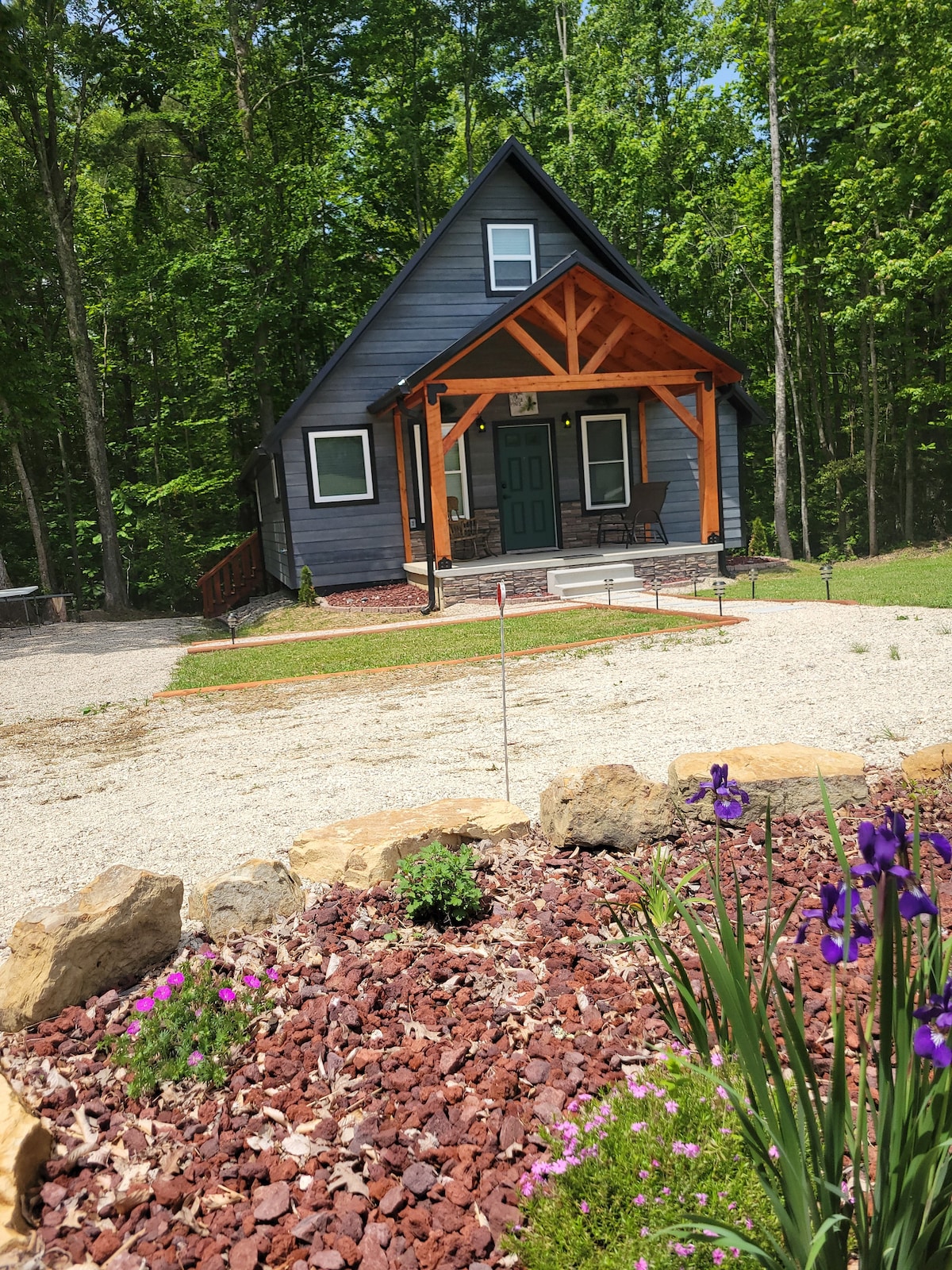 A cabin exterior is shown, featuring a dark-finished facade with wooden accents and a triangular roof. Stone landscaping with flowers and greenery is positioned in the foreground. A gravel driveway leads up to the cabin, surrounded by lush forest.