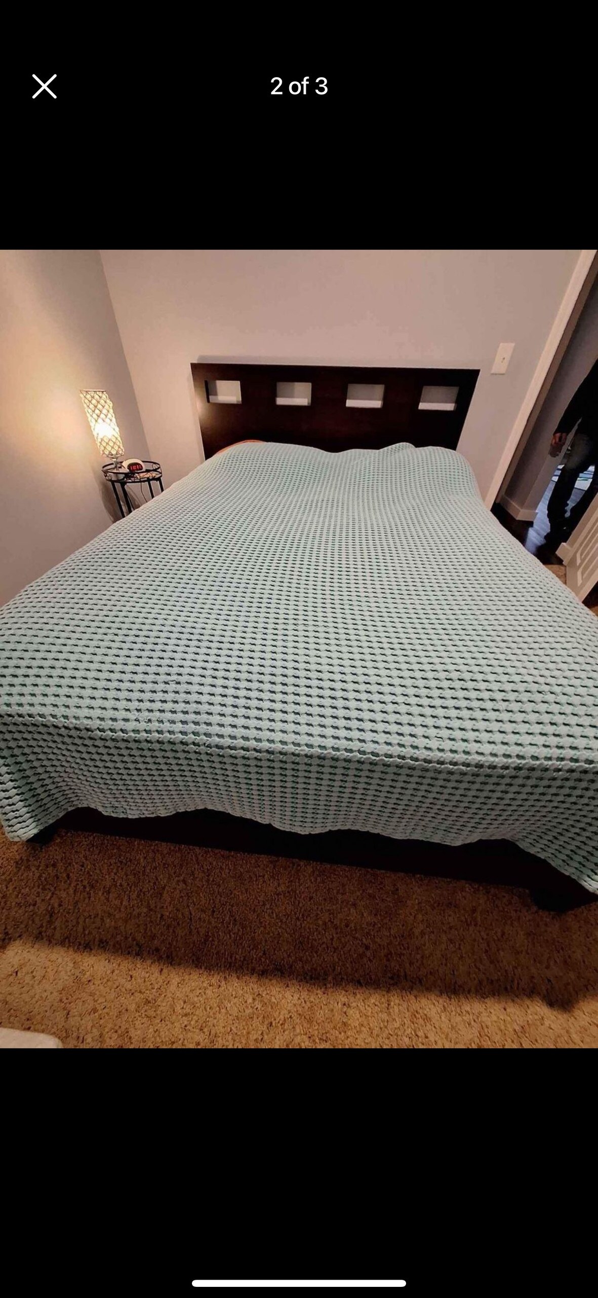 A spacious bed is covered with a textured green and white checkered blanket, creating a relaxed look. A softly illuminated lamp is positioned beside the bed on a dark nightstand, while a light grey wall provides a calming backdrop.