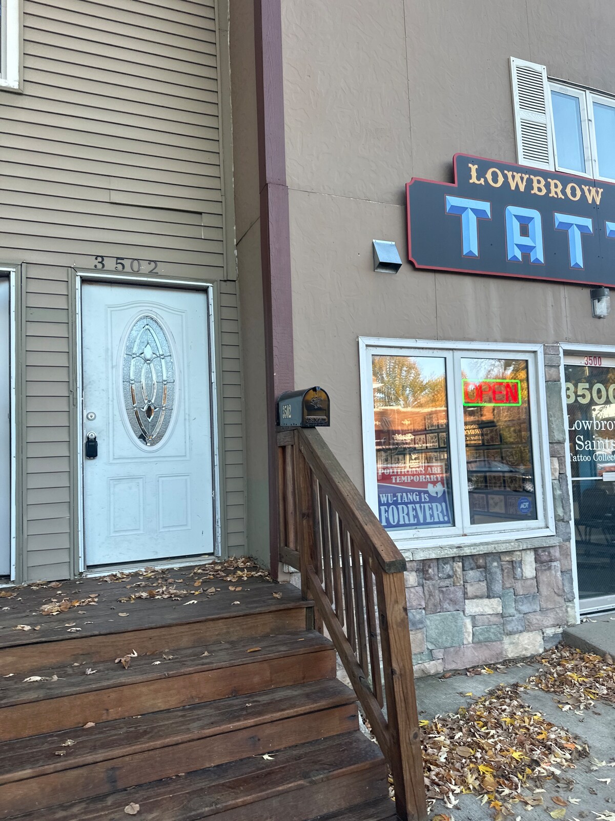 A front entrance is visible, featuring a decorative glass door with intricate designs. The steps leading to the door are constructed of wood, complemented by a small stone wall. Adjacent, a storefront displays a vibrant sign and large windows showcasing merchandise inside.