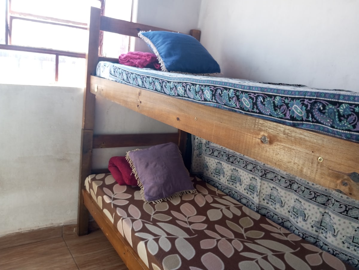 A cozy bunk bed is displayed, featuring a patterned mattress on the bottom and a colorful quilt draped over the top. Two pillows are placed atop the upper bunk, while additional cushions are positioned on the lower mattress. Natural light filters through a nearby window.