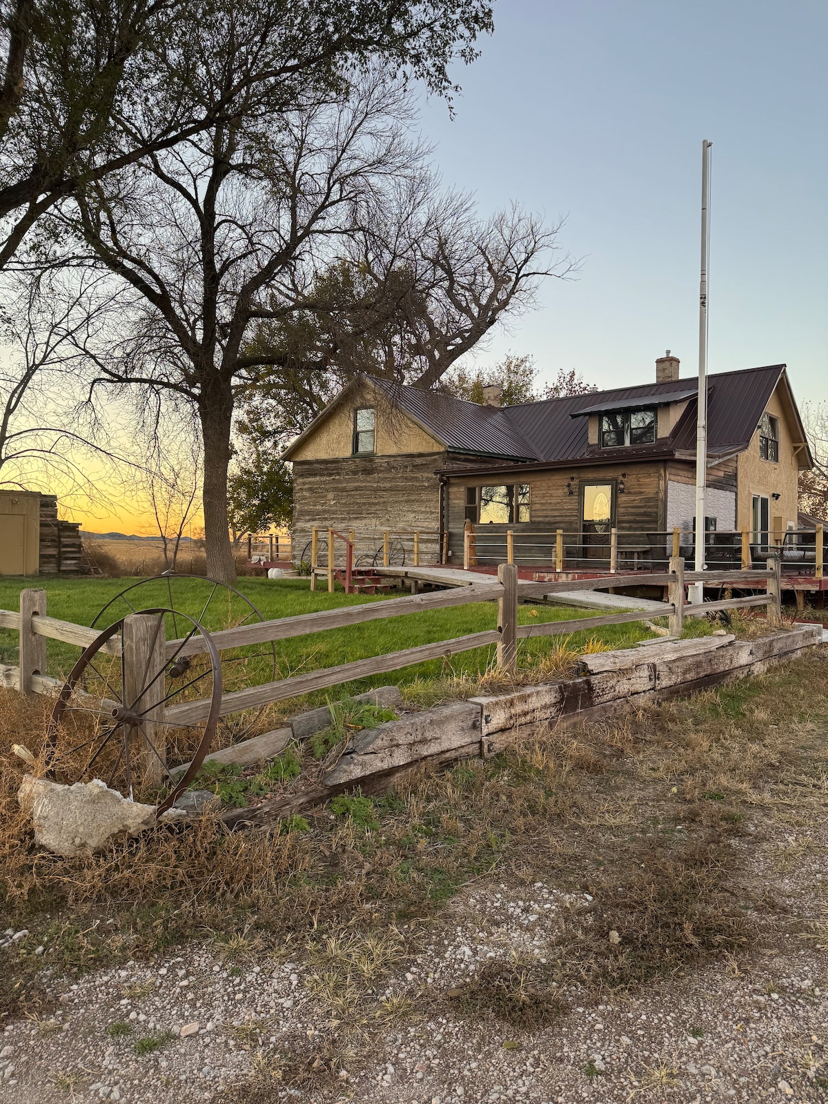 The exterior of a vintage house built from weathered wood is visible, surrounded by tall grass and a few trees. An old wagon wheel is positioned near a wooden fence, which lines the property. The sky presents a soft glow as the sun sets in the background.