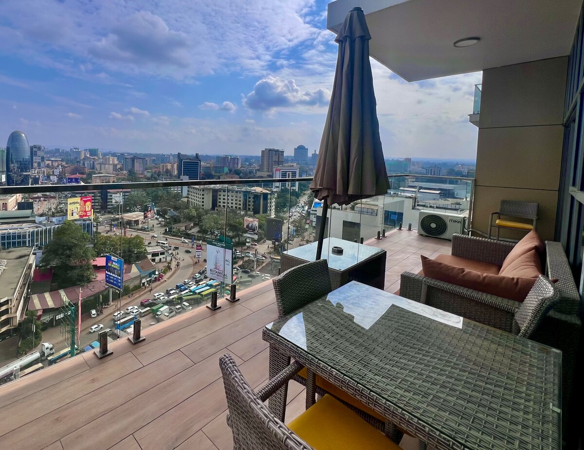 A spacious balcony offers stunning views of the Nairobi skyline, featuring a dining table and comfortable seating. An umbrella provides shade, while the cityscape below showcases a vibrant urban environment, seen from the 15th floor of the SkyNest building.