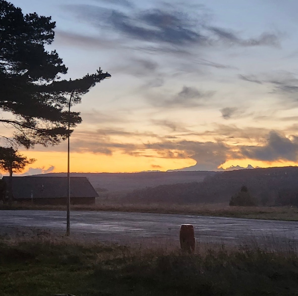A scenic view showcases a vibrant sunset sky with varying hues of orange and purple over a distant landscape. Silhouettes of trees and a small building are visible in the foreground against the softly illuminated clouds.