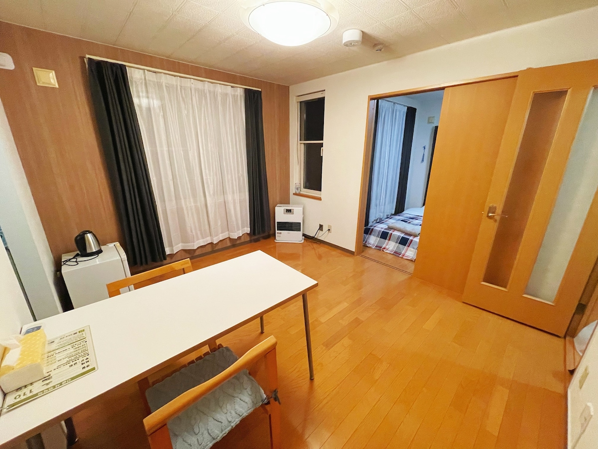 A functional living space is presented with a wooden floor and natural light filtering through sheer curtains. A simple table with two wooden chairs is positioned in the foreground, while an open door leads to a cozy sleeping area with a checkered blanket.