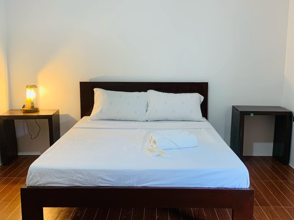 A simple and spacious sleeping area features a large bed with white linens and two neatly arranged towels. Soft lighting from a bedside lamp creates a gentle glow. Dark wooden side tables frame the bed, complementing the clean, minimalist design.
