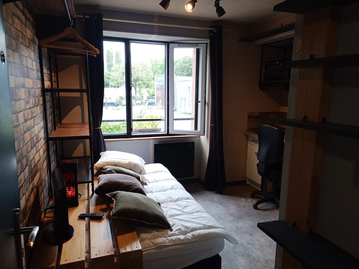 A studio space features a compact sleeping area with a spacious bed topped with crisp white bedding and decorative pillows. Large windows provide natural light, while a desk and chair are positioned nearby. Shelving units and a hanging rack add functional elements to the room.