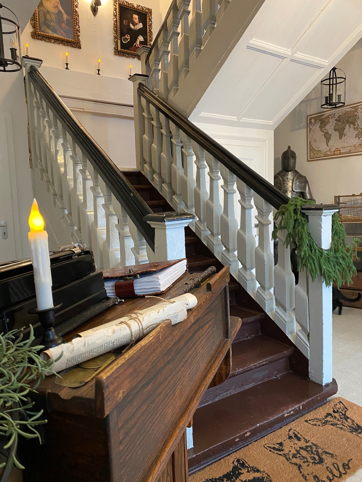 A staircase with a white railing ascends to the upper level, surrounded by framed portraits and decorative lighting. An antique table is positioned at the bottom, adorned with a candle and greenery. A vintage statue can be glimpsed in the background.