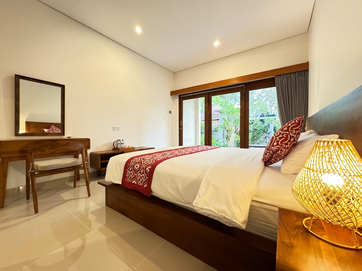 A cozy guest room features a spacious bed with crisp white linens and decorative pillows. A wooden desk and chair are situated nearby, with natural light filtering through sliding glass doors that open to a lush outdoor view. A wicker lamp provides gentle illumination.