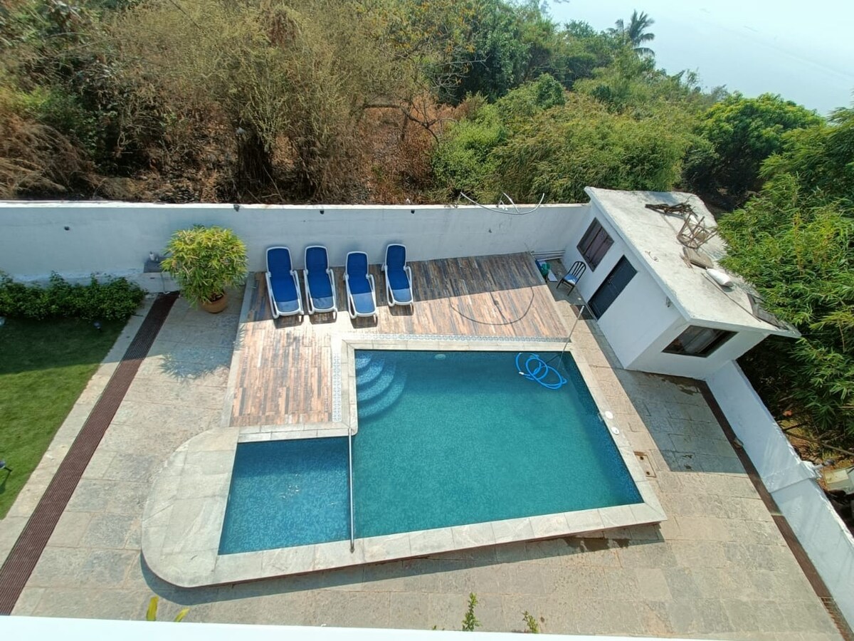 An aerial view of a pool area surrounded by lush greenery is depicted. A rectangular pool with clear water is bordered by natural stone. Six lounge chairs are arranged on a wooden deck, providing an inviting space for relaxation.