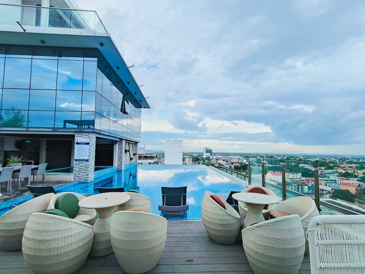The rooftop area offers a modern infinity pool with scenic views of the surrounding landscape. Comfortable lounge chairs are arranged around the pool, complemented by textured seating options. Floor-to-ceiling glass panels enhance the expansive feel of the space, welcoming natural light.