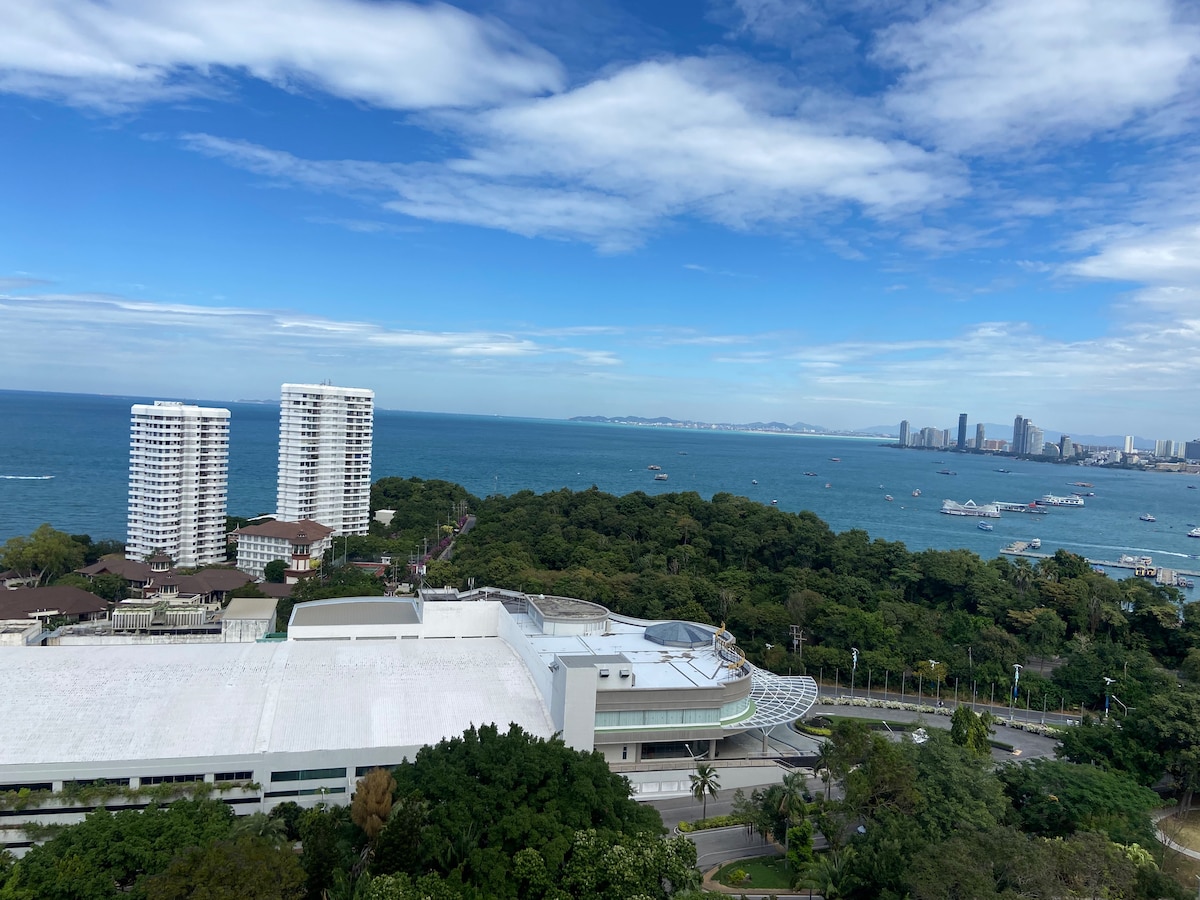 A panoramic view captures the Gulf of Thailand with a bustling coastline and distant city skyline. Towering white buildings are seen along the shore, surrounded by lush greenery, while boats dot the water, enhancing the serene coastal landscape.