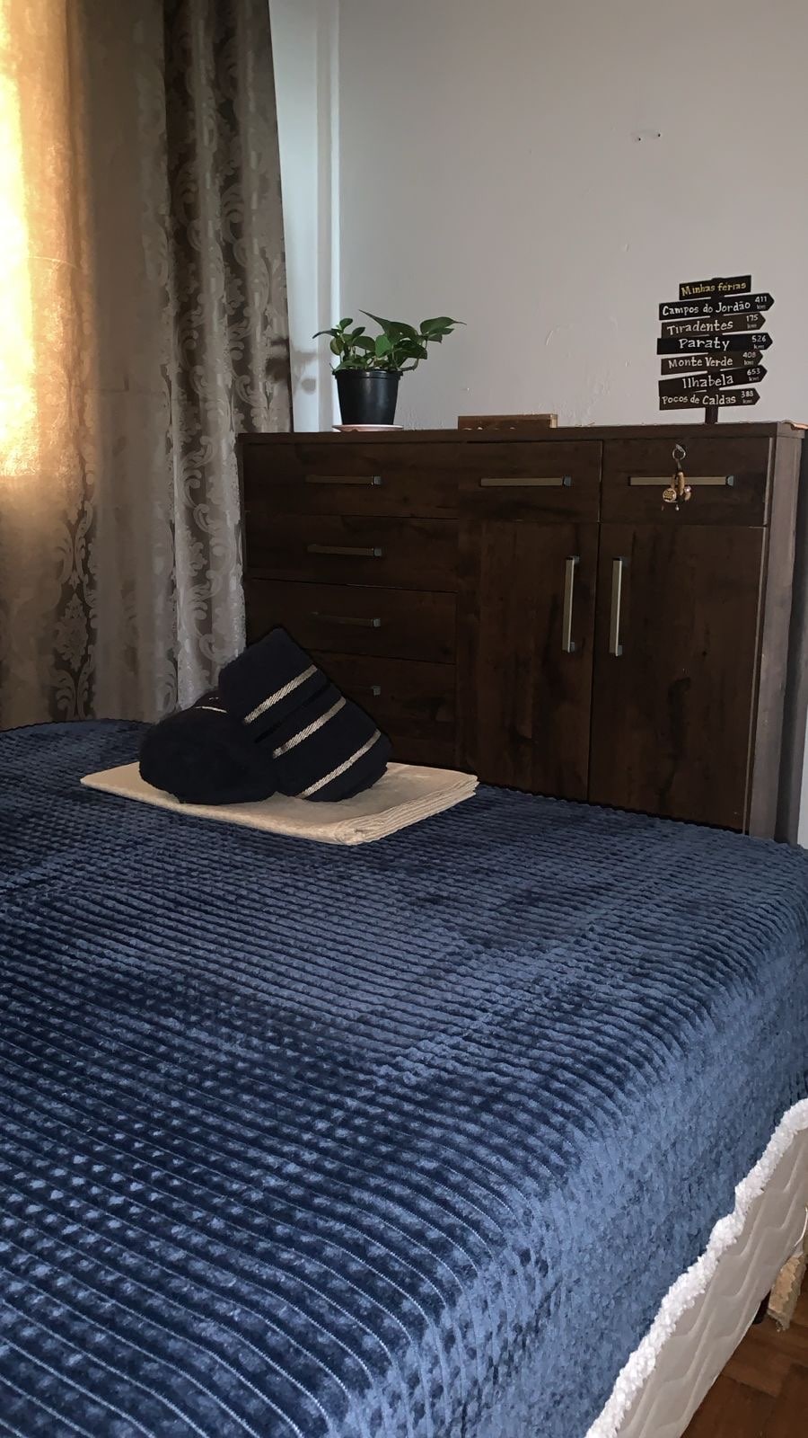 A serene sleeping space is showcased with a textured navy blanket draped over a spacious bed. An arrangement of black and white pillows rests at the head. A wooden dresser stands nearby, adorned with a small potted plant, while soft natural light filters through the window.