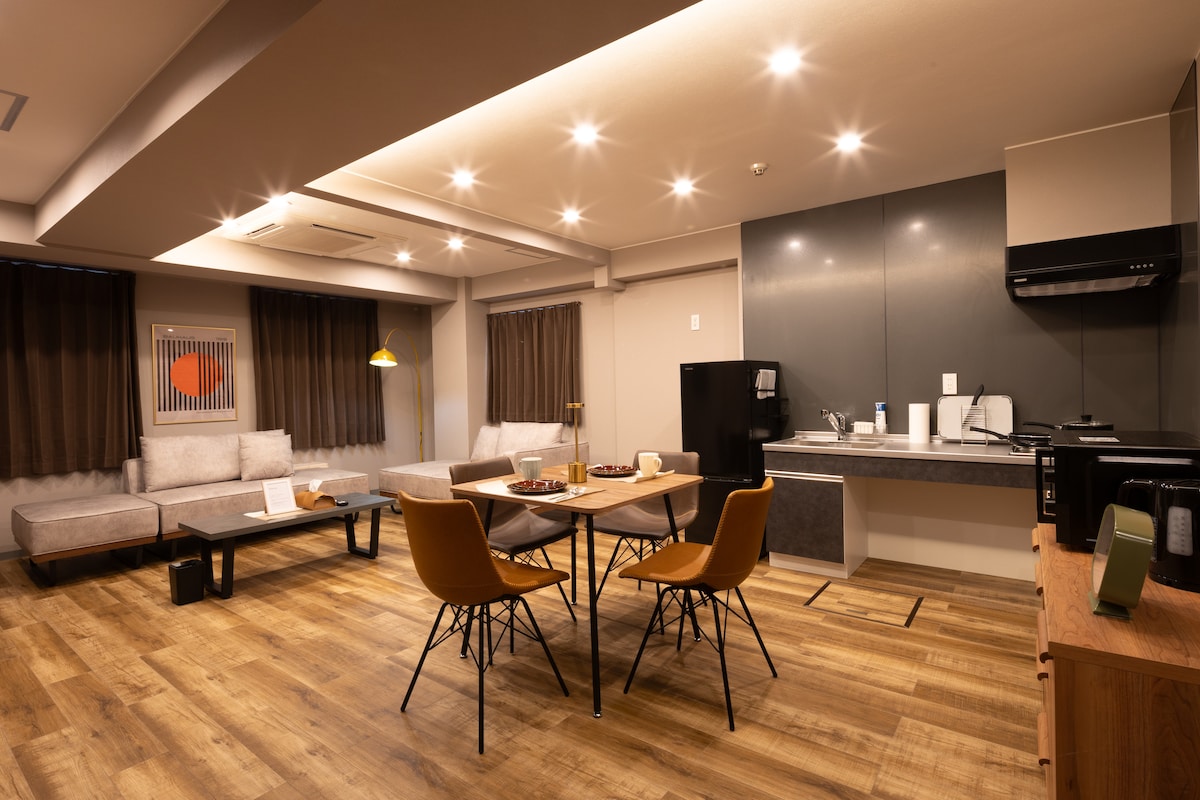 The spacious living area features a soft sectional sofa and a dining table with four chairs. A modern kitchenette is equipped with necessary appliances, including a refrigerator and microwave. Natural light filters through the brown curtains, creating a warm atmosphere.
