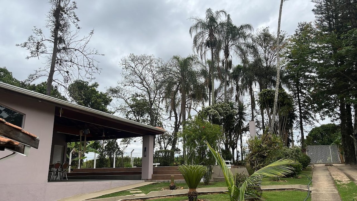 A spacious outdoor area is bordered by lush trees and palm plants. The property features a large covered patio that overlooks the greenery. The nearby pathway leads to additional outdoor spaces, providing a serene environment for relaxation.