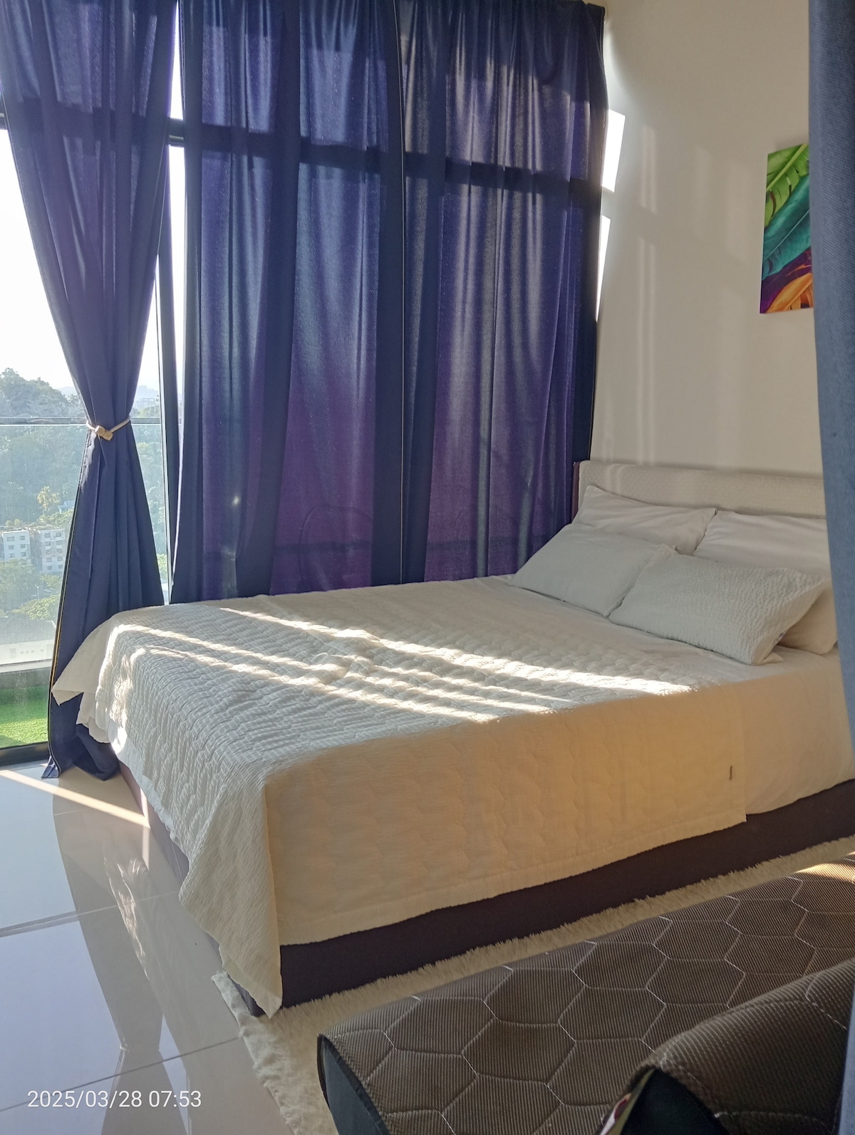 A well-lit sleeping area features a spacious bed with crisp white linens, positioned beside large windows covered with dark curtains. Natural light creates a serene ambiance, casting soft shadows across the floor. A colorful piece of wall art adds a touch of vibrancy to the neutral decor.