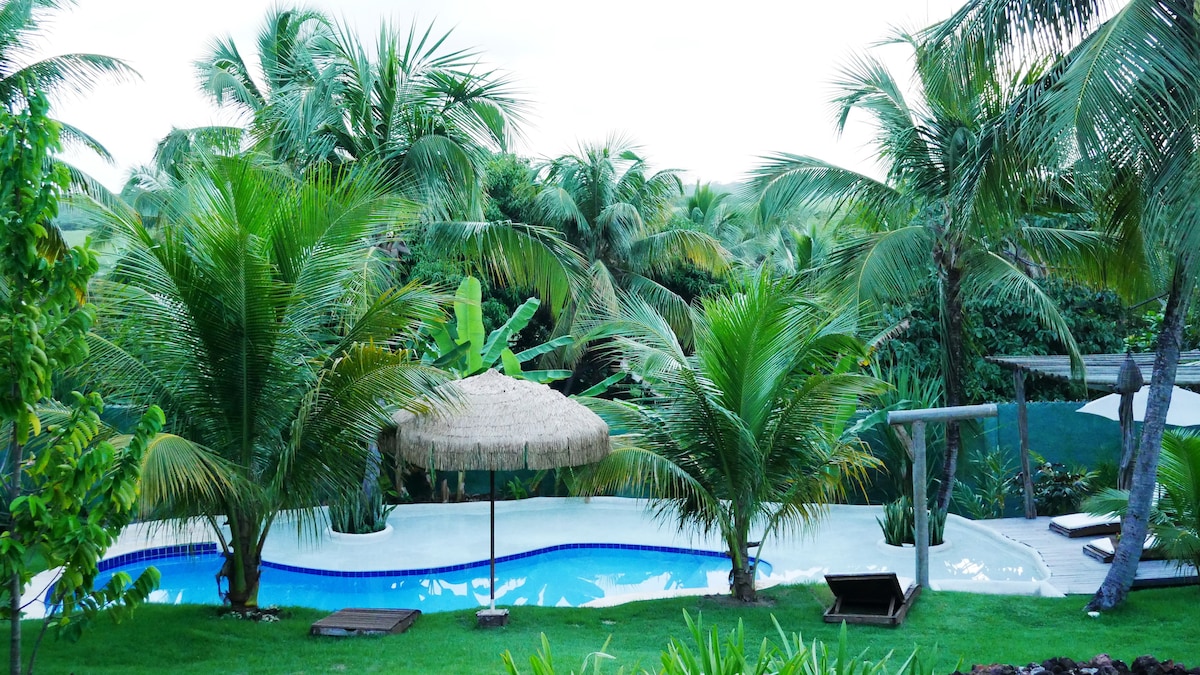 A serene outdoor area features a swimming pool surrounded by lush tropical plants. A thatched umbrella provides shade, and lounge chairs are positioned near the pool. The scene is complemented by palm trees framing the space, creating a tranquil atmosphere.