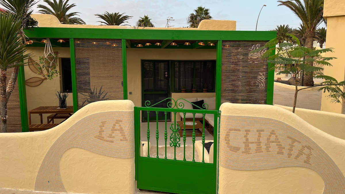 A vibrant green entry gate leads into a welcoming outdoor space adorned with palm trees. The area features a wooden table and chairs under a pergola, providing a shaded seating option. The front window reveals a cozy interior with decorative elements.