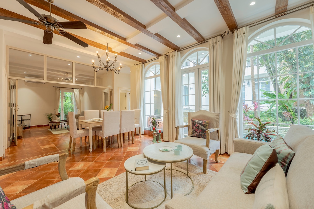 A spacious living area is highlighted by wooden beams and large windows, allowing natural light to fill the room. A comfortable sofa is paired with two coffee tables, while a dining table for six is visible in the background, surrounded by lush greenery.