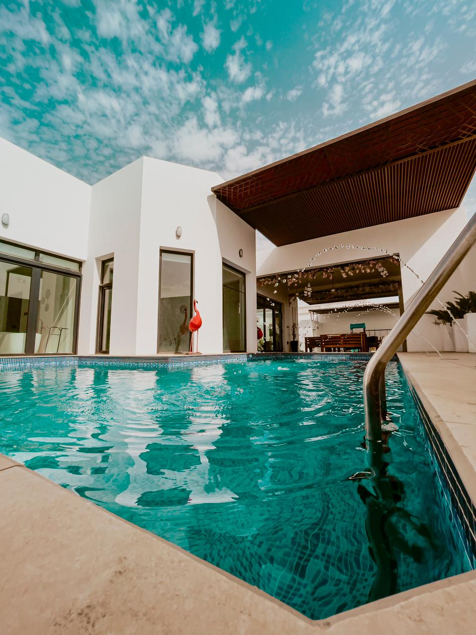 A tranquil swimming pool reflects the bright blue sky, with a modern architectural design surrounding it. Large glass doors provide access to the interior, while a shaded outdoor seating area is visible in the background, enhancing the space's relaxing atmosphere.