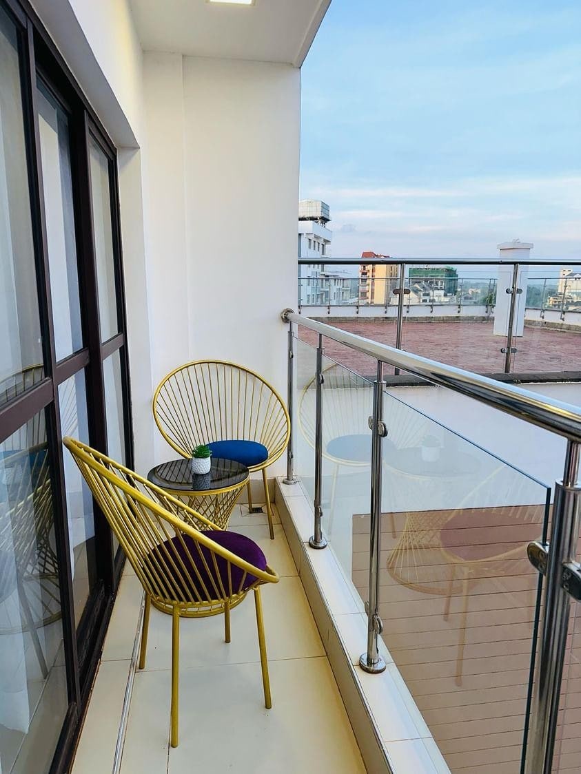 A spacious balcony is showcased, featuring two gold wire chairs with round cushions in purple and blue. A small table is positioned between the chairs. Glass barriers provide an unobstructed view of the surroundings, allowing natural light to illuminate the space.