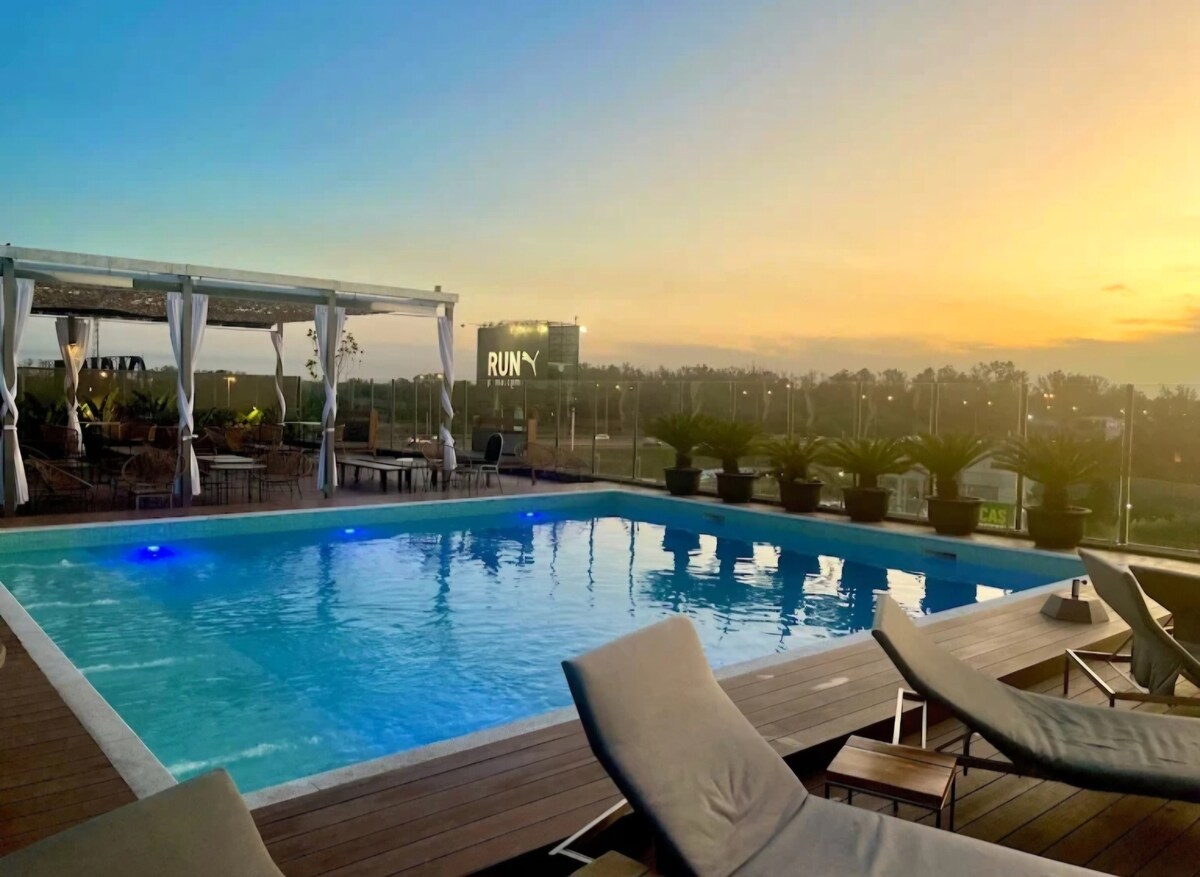 A rooftop pool area is highlighted at sunset, featuring a clear blue pool surrounded by sun loungers. Lush greenery and comfortable seating are visible, while a distant sign adds to the modern ambiance of the space.