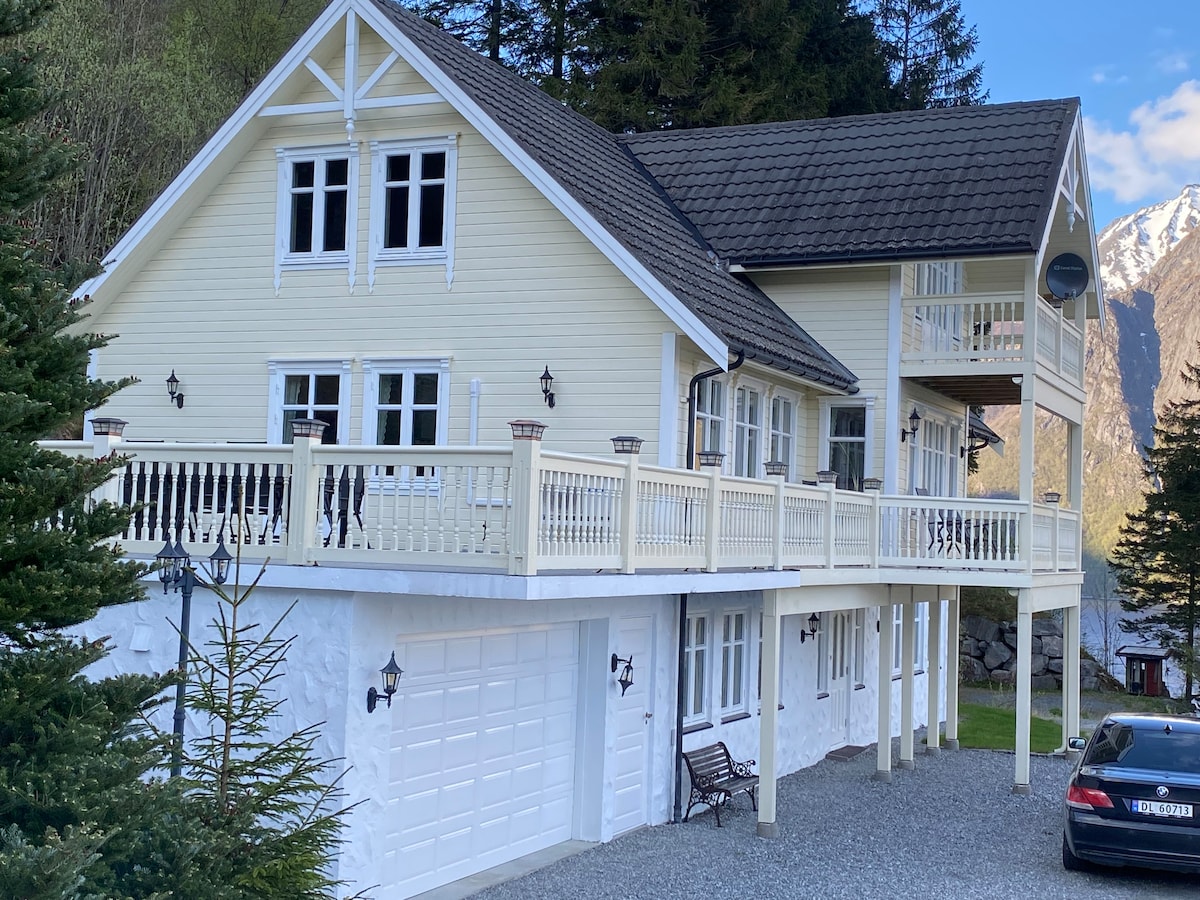 The exterior of a two-story holiday home is showcased, featuring light yellow siding and a steeply pitched roof. A spacious balcony surrounds the upper level, while a garage is situated below. The property is set against a backdrop of lush greenery and mountains.