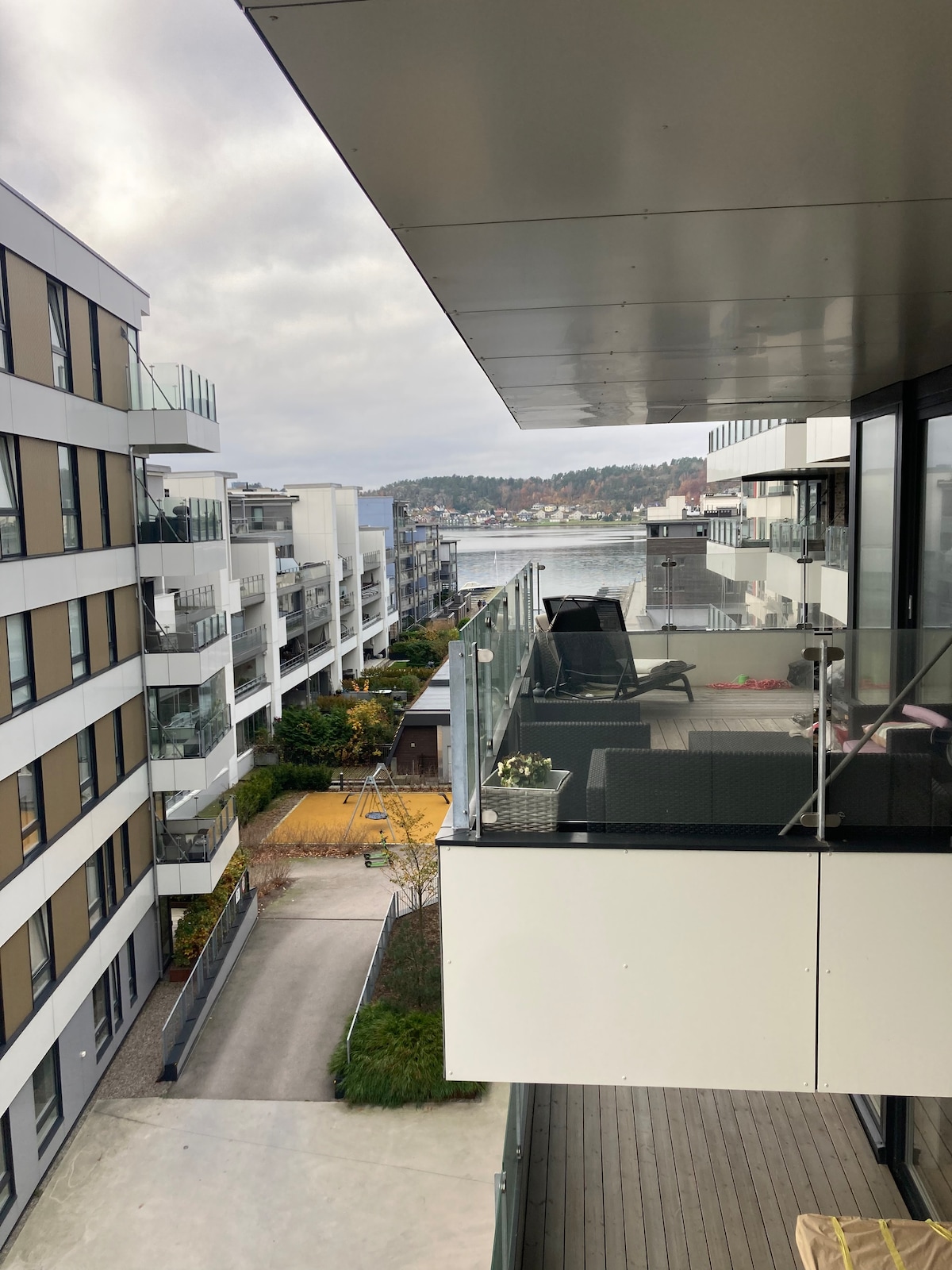 A spacious terrace is visible, featuring outdoor seating and a view of the nearby waterfront. The surrounding buildings exhibit modern architecture with large glass windows. Lush greenery is present in the courtyard areas, enhancing the serene environment.