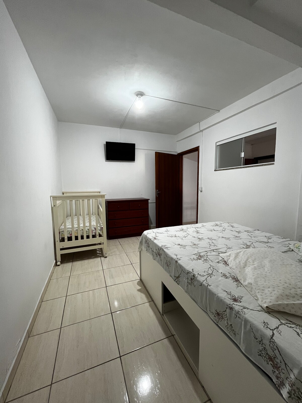 A simple and functional bedroom features light tile flooring and neutral white walls. A spacious bed with linens is positioned beside a baby crib. A dresser stands against the wall, and a small wall-mounted TV is visible. Natural light enters through a window with a frosted glass panel.