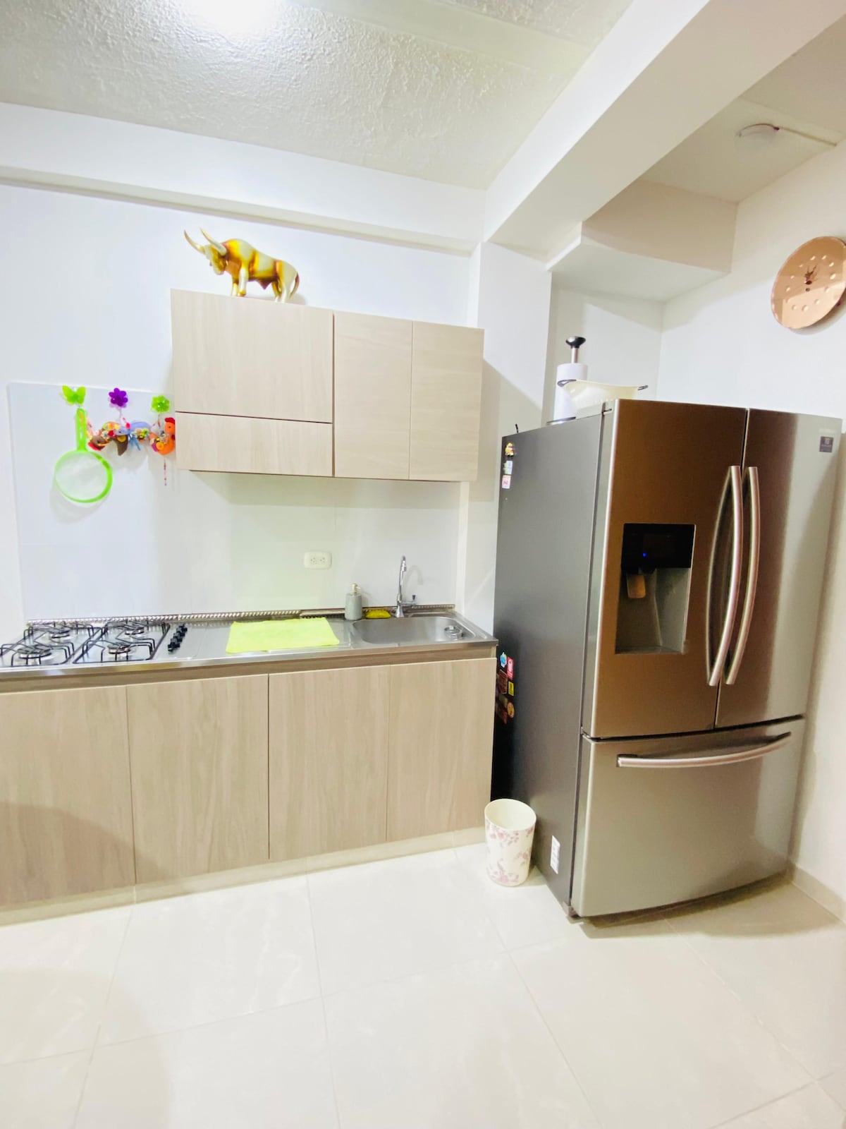 A modern kitchen is presented with light wood cabinetry and a stainless steel refrigerator. A gas stove is integrated into the countertop, accompanied by a sink. Decorative elements, including colorful utensils and a small sculpture, add a touch of charm to the functional space.
