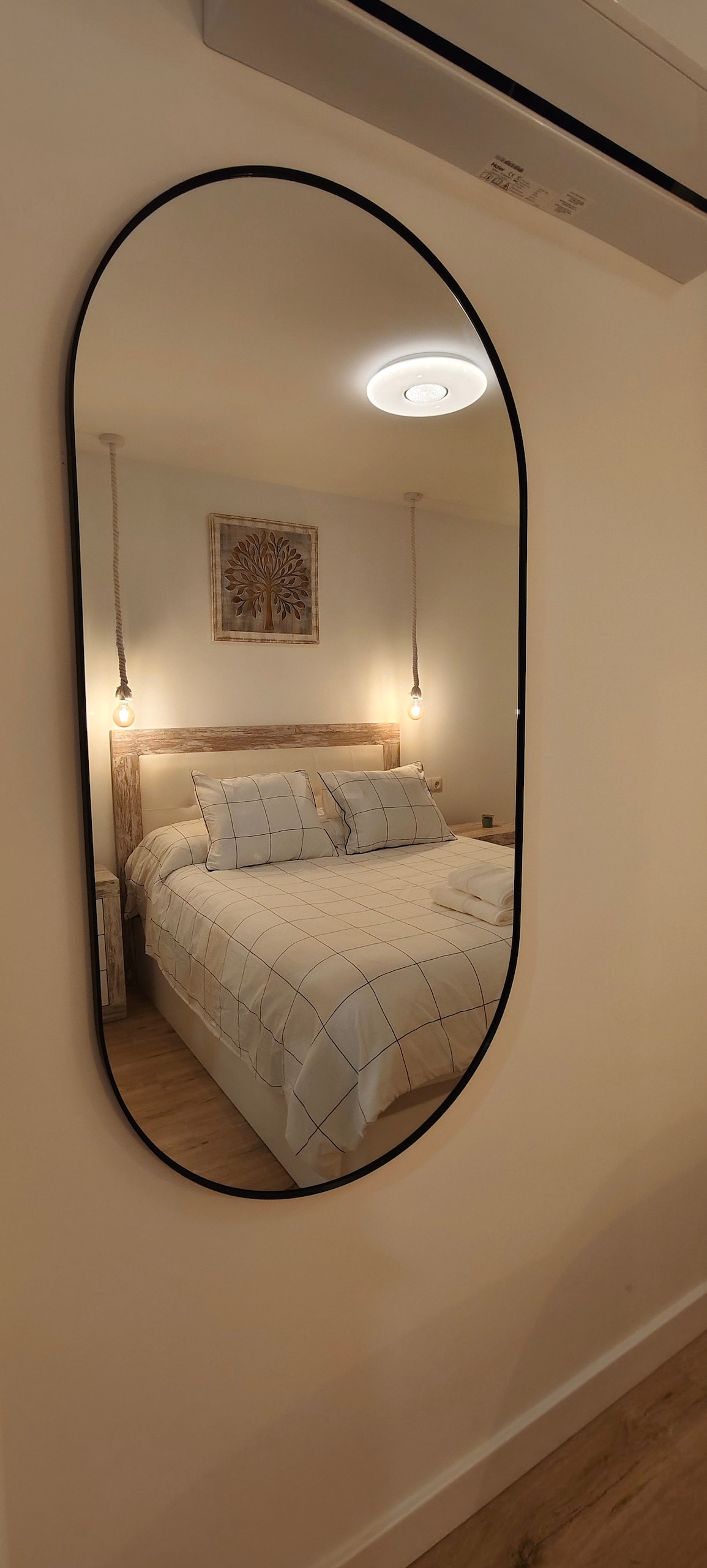 A spacious bedroom is reflected in an oval mirror. The bed features a light-colored, checkered duvet and cushions, with soft lighting provided by pendant bulbs. A decorative piece is hung above the headboard, and a neutral wall complements the warm tones of the space.