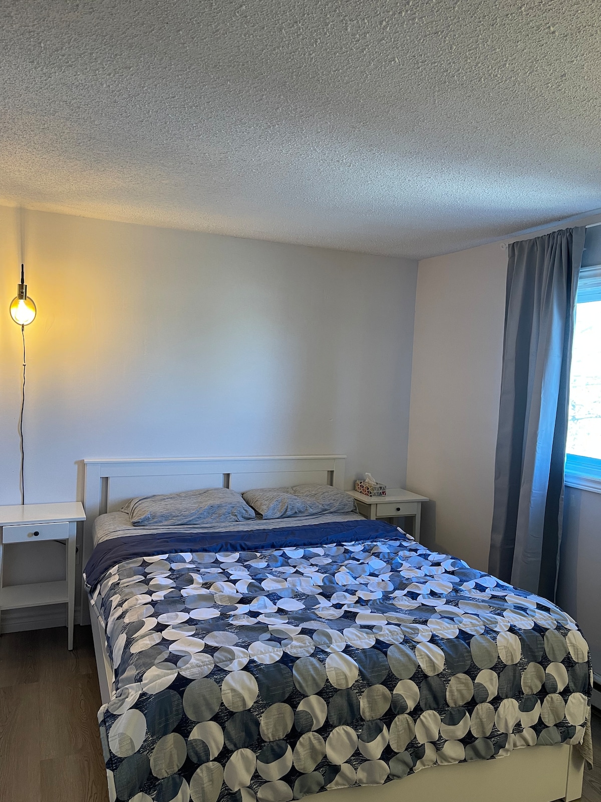 A neatly arranged bedroom features a spacious bed with a patterned blue and white blanket. Two nightstands are placed beside the bed, and a pendant light provides soft illumination. A window with sheer curtains allows natural light to brighten the room, enhancing its cozy atmosphere.
