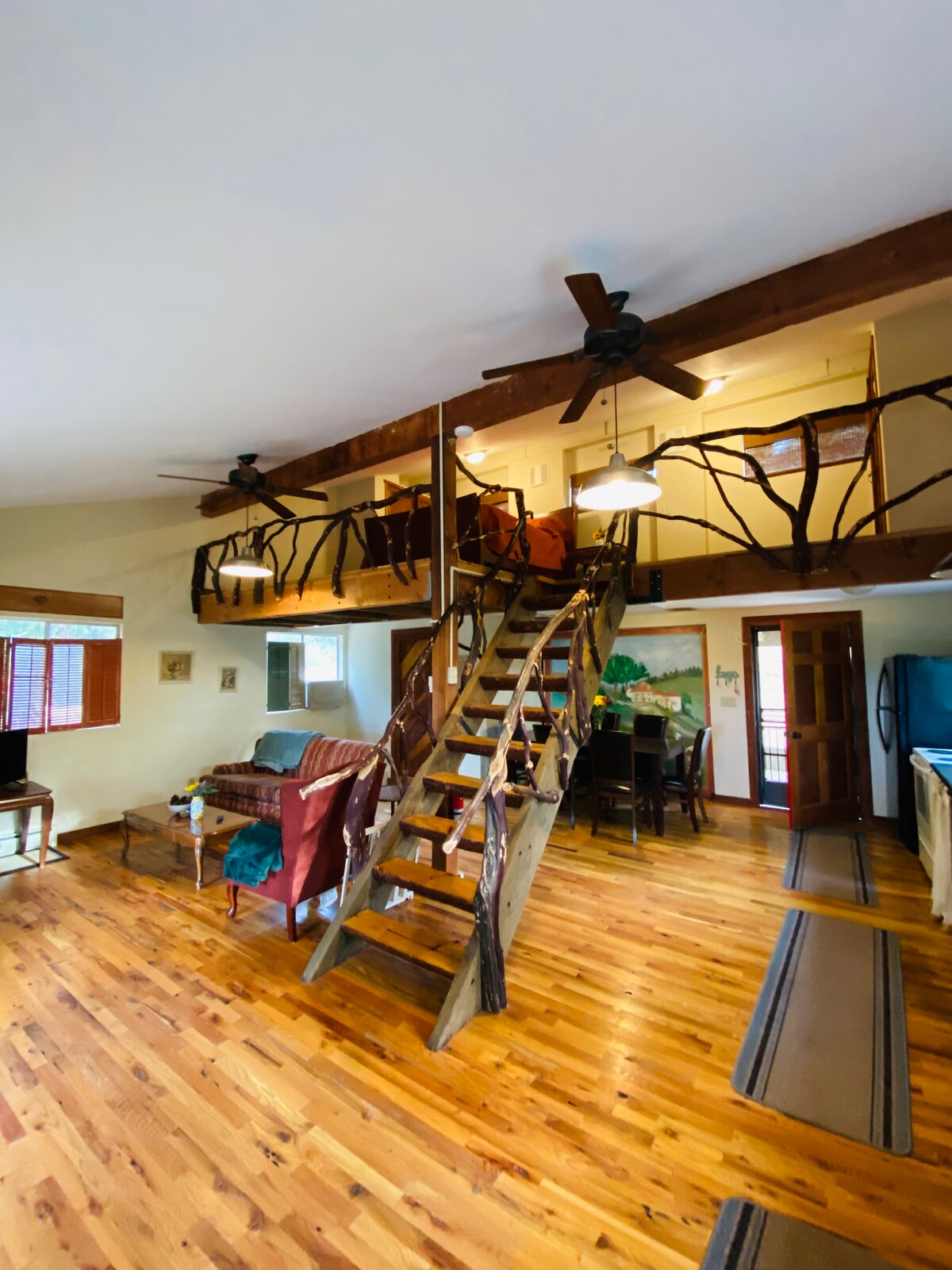 An open floor plan is featured, showcasing wooden floors and rustic wooden beams. A staircase leads to a loft area, while a dining table and seating areas are visible. Natural light enters through several windows, creating an inviting, spacious environment.