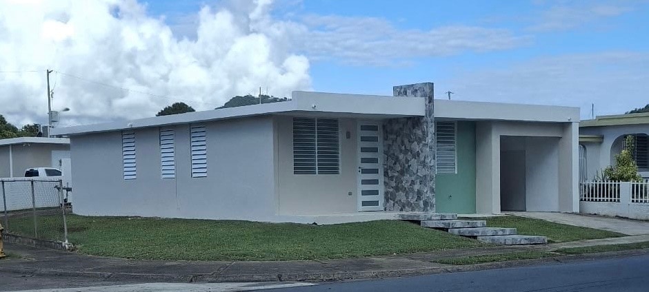 A modern one-story house features a light-colored exterior with horizontal and vertical window styles. A stone accent wall is noticeable near the front entrance. A well-manicured lawn provides a spacious area in front of the house, while a concrete pathway leads to the entrance.