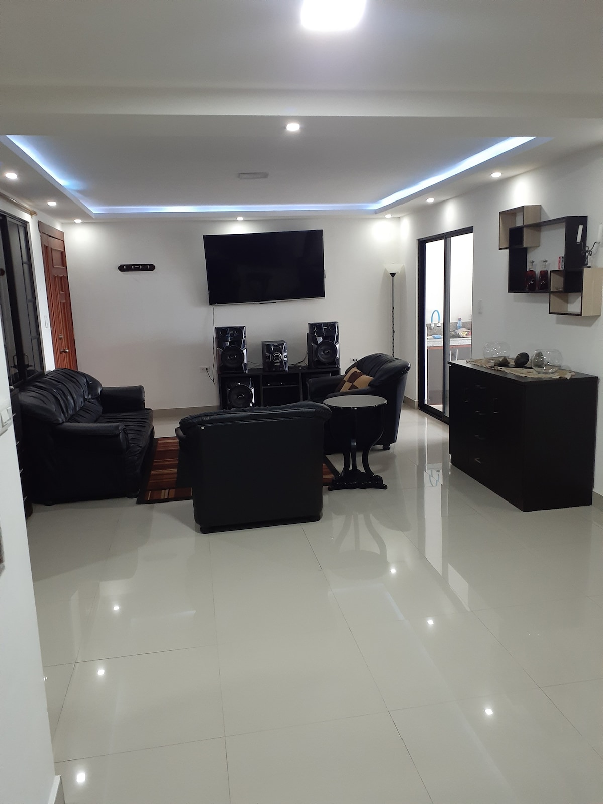 A modern living area is featured with sleek leather seating arranged around a central rug. A wall-mounted television is positioned above a black entertainment unit, complemented by built-in speakers. Natural light enters through large windows, contributing to the spacious feel of the room.
