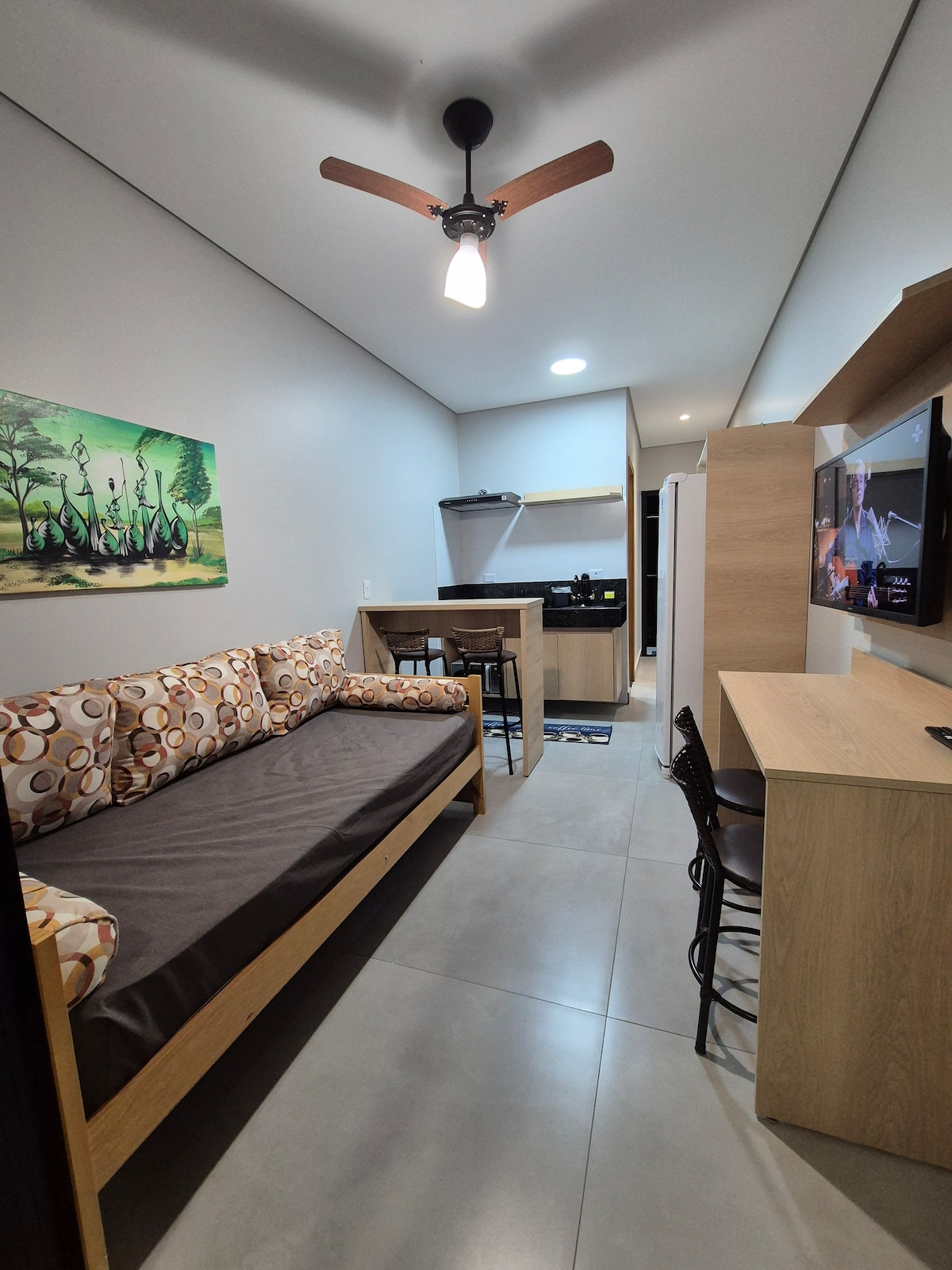 An open living area is presented with a modern design featuring a cozy sofa lined with circular-patterned cushions. A dining table and chairs occupy one side, while a kitchenette is partially visible, equipped with essential appliances. A wall-mounted TV is positioned above a light wood console.