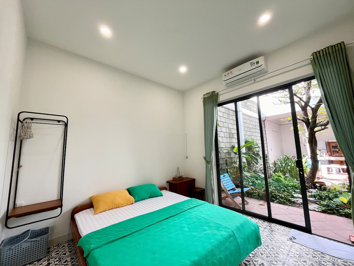 A bright and airy room features a spacious bed with a green blanket and yellow accent pillow. Large glass doors open to a garden view, while light-colored walls and patterned flooring enhance the space. A nightstand sits beside the bed, and a simple clothing rack is present.