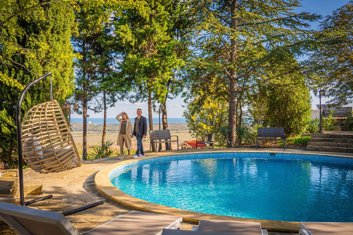 A serene outdoor area features a circular swimming pool surrounded by sun loungers and trees. A decorative hanging chair adds an inviting touch. In the background, two people stroll while enjoying the scenic view of the landscape, enhancing the tranquil ambiance.