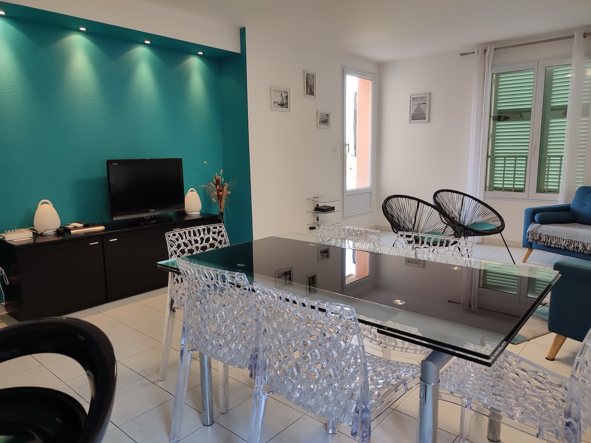 A light-filled living area features a modern glass dining table surrounded by transparent chairs. A plush blue sofa is positioned nearby, and a television is mounted on the wall. The decor includes subtle accents of color on the walls and framed artwork.