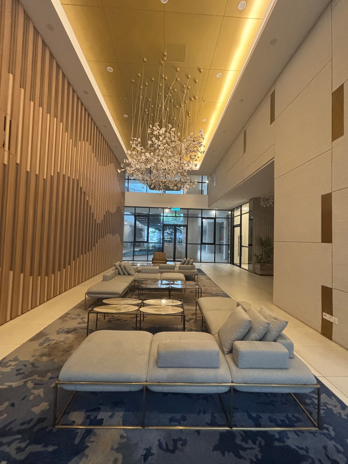 A modern lobby area features a combination of wood paneling and elegant lighting. Comfortable seating is arranged around glass-top tables on a decorative rug, with a large chandelier hanging overhead. Floor-to-ceiling windows provide natural light, creating an inviting space.