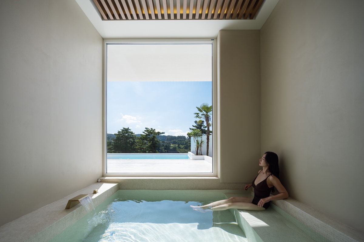 An indoor spa area is shown with a clear window providing a view of the outdoor pool and surrounding landscape. Soft light filters through, highlighting the serene water in the jacuzzi and the minimalist design of the space.