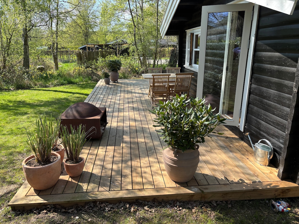 A spacious wooden terrace extends from the main house, featuring a dining table and chairs. Potted plants are arranged around the area, adding a touch of greenery. Natural light bathes the space, surrounded by trees and a peaceful outdoor setting.