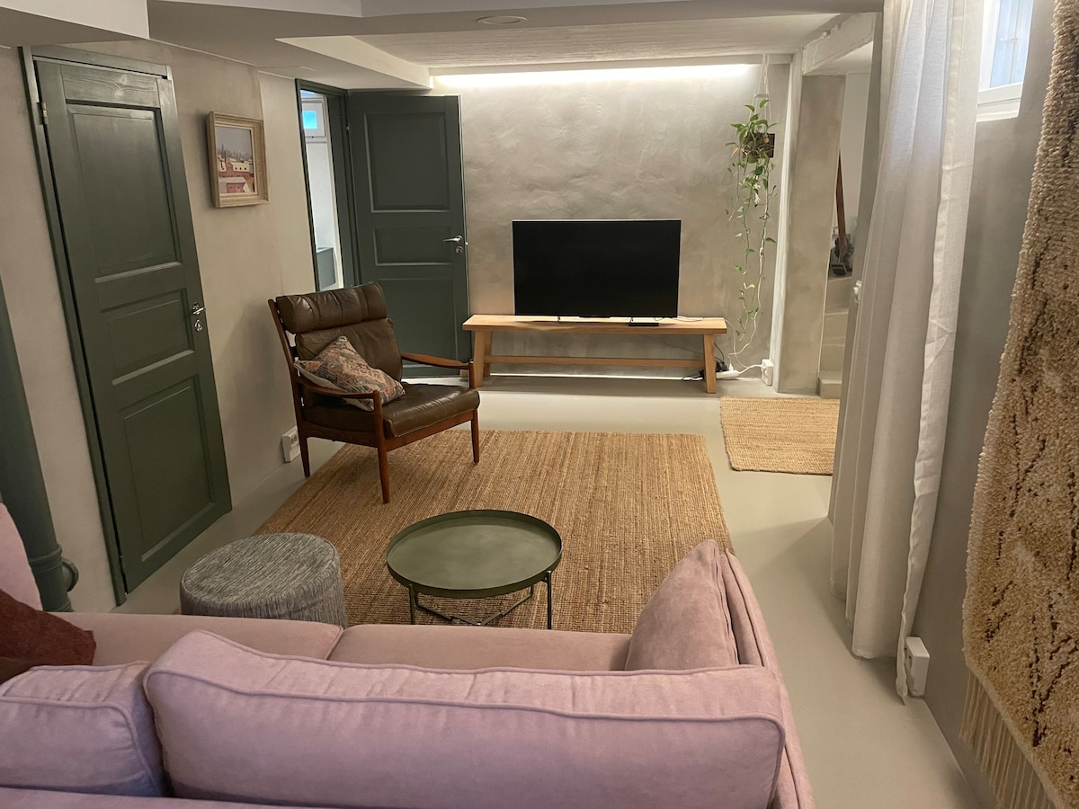 A cozy living area is furnished with a pastel-colored sofa and a brown leather chair. A green coffee table is placed on a woven rug, with a wall-mounted television situated on a natural wood stand. Soft lighting accentuates the neutral tones of the room.