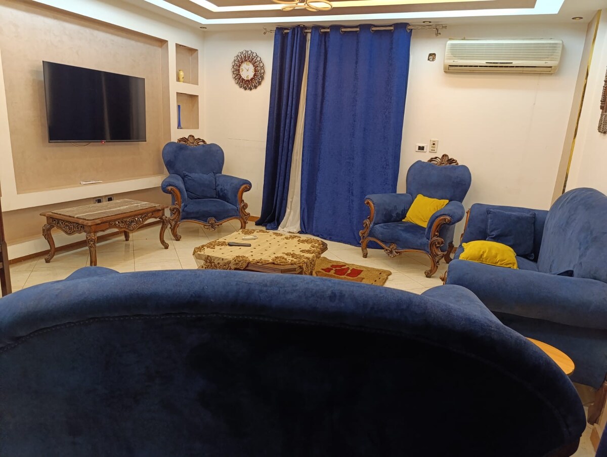A cozy living area features two blue upholstered chairs with ornate wooden accents. A low, decorative coffee table rests at the center, surrounded by a welcoming atmosphere. A large television is mounted on the wall, and deep blue curtains frame the space, allowing for privacy and comfort.