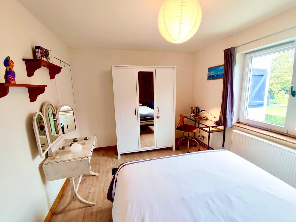 A well-lit room features a spacious bed with a light-colored duvet. A white wardrobe stands against one wall, while a small desk and chair are positioned nearby. Three mirrors are mounted on a shelf, and a window offers a view of the garden.