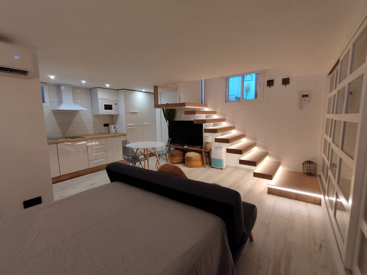 A welcoming studio space features a cozy sleeping area with a spacious bed. An open kitchen layout includes modern appliances. A staircase with illuminated steps leads to a comfortable seating area with a TV. Soft lighting enhances the tranquil indoor ambiance.
